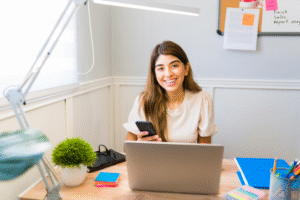 A smiling virtual assistant working on her laptop while holding her smartphone.