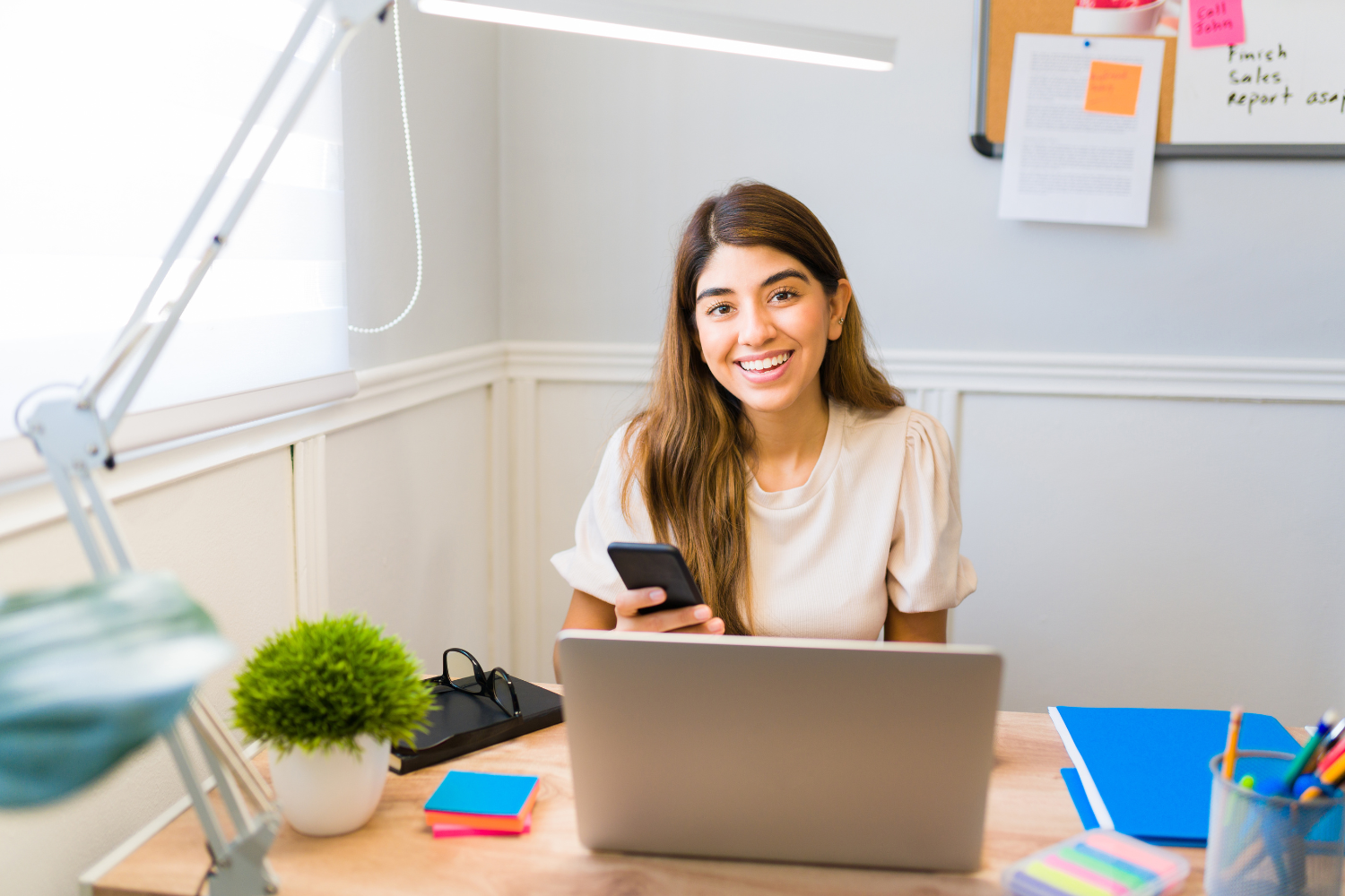 A smiling virtual assistant working on her laptop while holding her smartphone.